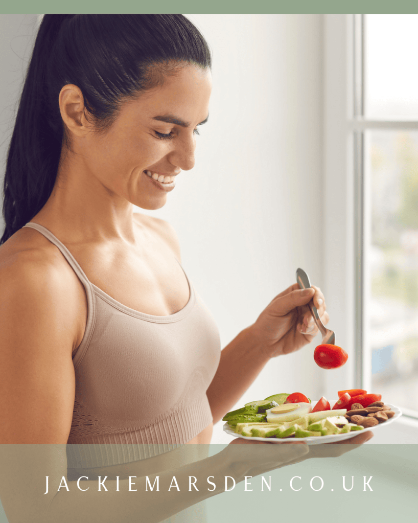 A smiling woman in a fitted tank top holds a plate of fresh vegetables and nuts, looking content while preparing to eat.